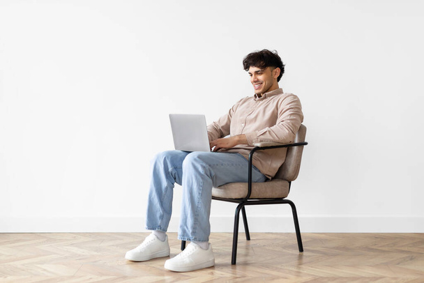 Homme indépendant moyen-oriental prospère avec ordinateur portable travaillant en ligne tout en étant assis dans une chaise sur fond blanc à la maison, beau mec arabe utilisant l'ordinateur, naviguant sur Internet pour le travail et l'étude - Photo, image