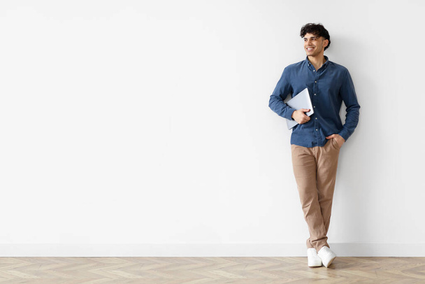 Młody Bliskowschodni facet pozował trzymając PC Laptop stojący w białym studio Wall Indoors, Full Length Shot. Człowiek z komputerem patrzący na przestrzeń kopiowania, Reklama oferty online - Zdjęcie, obraz