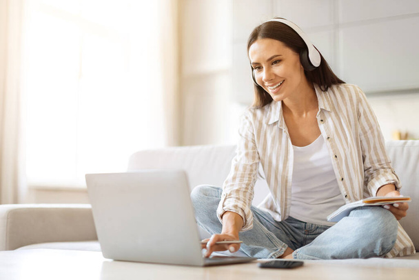 Vzdálené učení. Usmívající se mladá žena v bezdrátových sluchátkách Studium s notebookem doma, šťastný evropský žena pomocí počítače pro on-line vzdělávání, zatímco sedí na gauči v obývacím pokoji - Fotografie, Obrázek