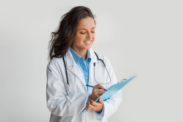 Skupiona lekarka w białym płaszczu, trzymająca schowek i robiąca notatki, oferująca łagodny uśmiech, odzwierciedlający profesjonalizm i troskę w środowisku medycznym - Zdjęcie, obraz