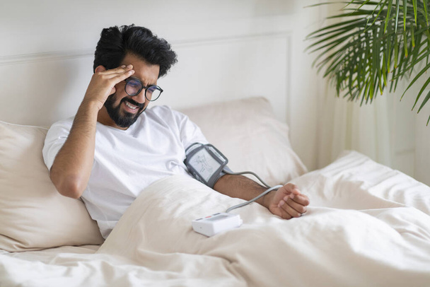 Hypertension Concept. Young Indian Man Checking Blood Pressure With Upper Arm Monitor While Sitting In Bed, Sick Eastern Male Measuring Arterial Tension And Touching Head, Having Acute Headache - Photo, Image