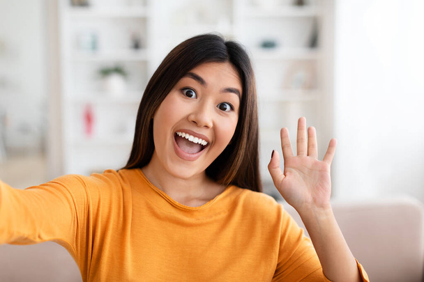 Pozytywnie piękna młoda azjatycka kobieta w pomarańczowym rękawie robi selfie w domu, radośnie macha i uśmiecha się do kamery, influencer pokazuje swoje nowe mieszkanie. Koncepcja blogowania - Zdjęcie, obraz