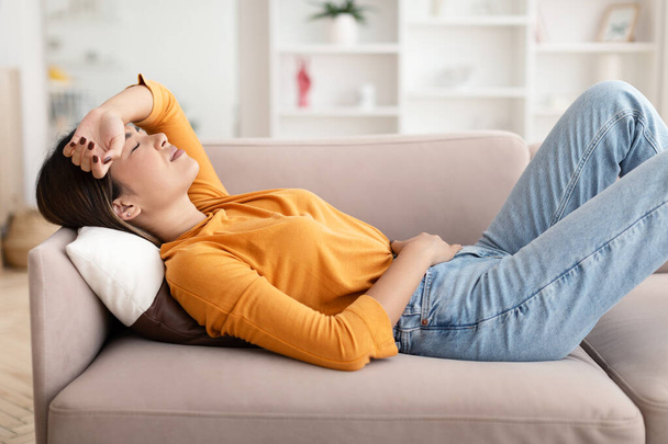 Feeling unwell, menstruation, flu, migraines and illness. Sad despair young asian woman lying on couch suffering from headache and abdominal pain in living room home interior - Foto, Bild