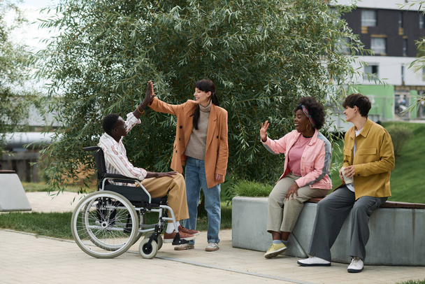 Grupo de amigos felices de ver a su amigo con discapacidad mientras está sentado en el banco en el parque - Foto, imagen