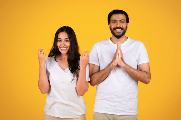 Usmívající se mladý arabský pár zkřížené prsty, aby se modlit gesto, izolované na žlutém pozadí studia. Naděje, dobré zprávy, sny se plní a velké přání zvítězit, emoce lidí - Fotografie, Obrázek