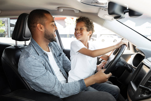 父親 親権 家族 愛情。 愛する黒人の若い父親と19歳の息子が一緒に車を運転しています. 子供はパパのラップに座って,ステアリングホイールで手を握り,彼のパパとつながる - 写真・画像
