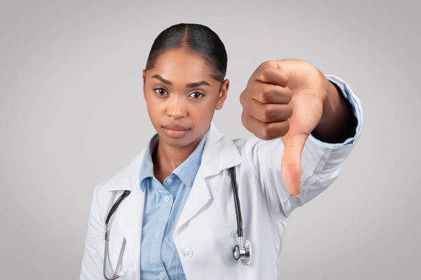Sad adult african american woman doctor in white coat make thumb down sign with hand, isolated on gray background, studio. Medical help, bad news gesture, negative review - Photo, Image