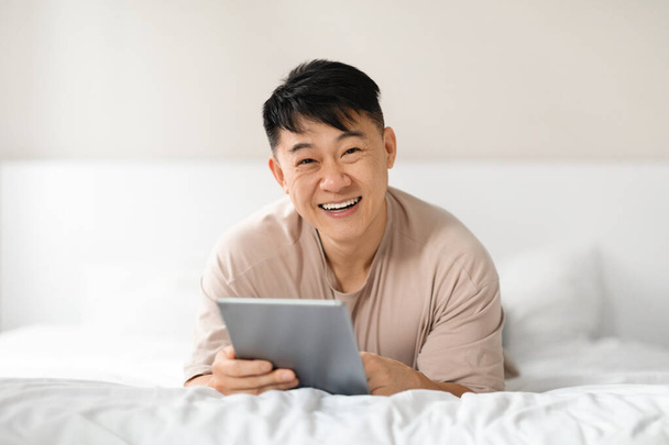 Rilassato felice uomo asiatico maturo in casa rilassarsi a letto dopo essersi svegliati al mattino, utilizzando tablet digitale e sorridente, scorrendo sui social media, websurf, lettura di notizie, spazio copia - Foto, immagini