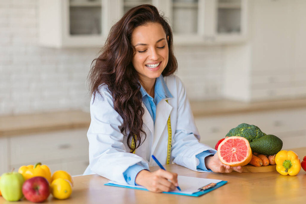 Улыбающийся диетолог в лабораторном халате пишет на планшете, демонстрируя нарезанный грейпфрут и ассорти свежих фруктов и овощей на деревянном столе в помещении - Фото, изображение