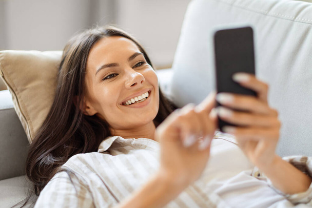 Młoda uśmiechnięta piękna kobieta ze smartfonem spoczywa na kanapie w domu, Happy Millennial Kobieta leży na kanapie i przy użyciu telefonu komórkowego, wiadomości z przyjaciółmi lub przeglądanie nowej aplikacji na zakupy - Zdjęcie, obraz