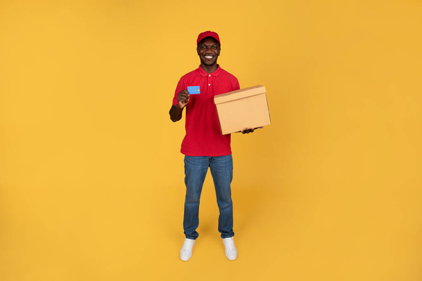 笑顔のミレニアルブラック男宅急使のホールドボックスは,オレンジ色のスタジオの背景に隔離された支払いのためのクレジットカードをお勧めします. ホーム 配達,仕事およびサービス,お金および財政,全長 - 写真・画像