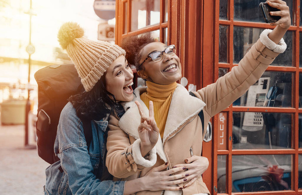 kaksi ystävää, tyttöystävää ja naista, jotka käyttävät matkapuhelinta, kameraa ja ottavat selfien punaiseen puhelinkoppiin Englannin kaupungissa.Travel Lifestyle käsite - Valokuva, kuva