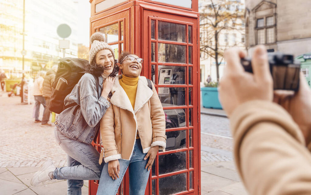 δύο φίλοι, φίλη και γυναίκες που χρησιμοποιούν ένα κινητό τηλέφωνο, κάμερα και τη λήψη selfie κατά ένα κόκκινο phonebox στην πόλη της Αγγλίας. Travel Lifestyle έννοια - Φωτογραφία, εικόνα