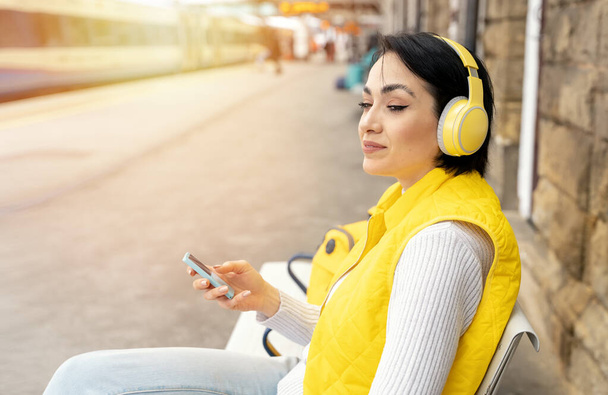 Young woman listening to music via headphones at railway station Travel with music Enjoying travel concept - Photo, Image