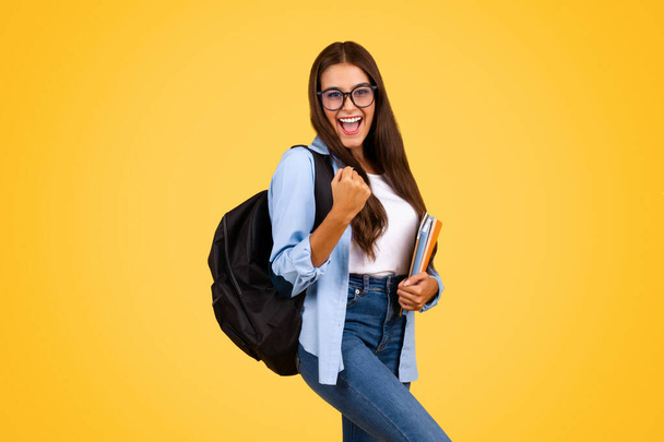Joven estudiante alegre bastante inteligente con mochila en gafas, disfrutar del éxito, aislado en el fondo del estudio amarillo. Educación y estudio, estilo de vida en la escuela, emociones, diversión - Foto, imagen