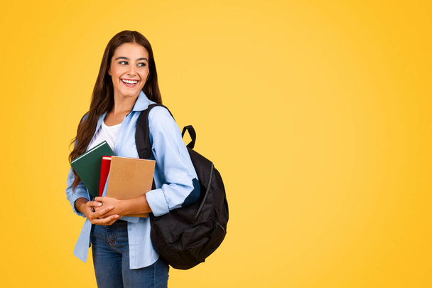 Positive pretty young student lady with backpack and books look at copy space, isolating on yellow studio background. Education, study, lifestyle at school, ad and offer - Photo, Image