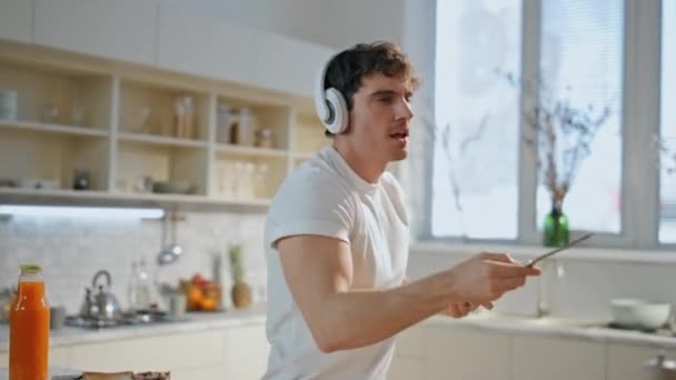 Chlápek, co poslouchá hudbu v kuchyni a má na sobě bezdrátová sluchátka. Radostně uvolněný muž tančí ve sluchátkách a připravuje chutné tousty k snídani v útulném interiéru kuchyně. Melomaniak vaření ve sluchátkách - Záběry, video