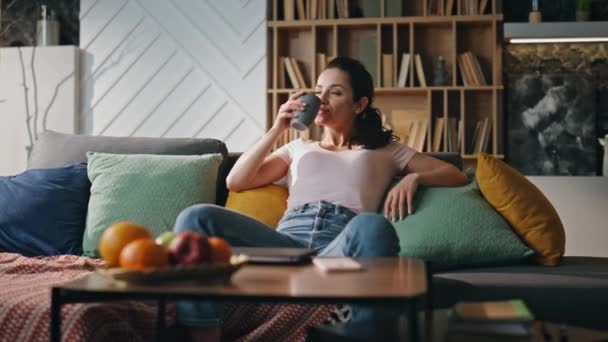 Relaxed woman freelancer drinking tea sitting cozy couch living room alone. Calm happy businesswoman relaxing after remote work in apartment. Attractive brunette enjoying tasty beverage home evening. - Footage, Video