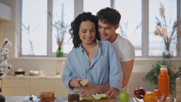 Happy lovers cooking at home hugging enjoy tenderness closeup. Loving husband cuddling laughing wife cutting apple on kitchen table. Married couple have fun preparing romantic breakfast lunch together - Footage, Video