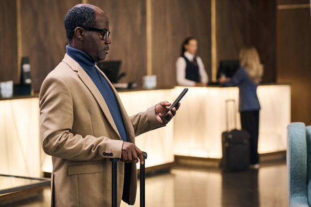 Αφροαμερικανός επιχειρηματίας ελέγχει τη διαμονή του online στο smartphone, ενώ στέκεται με τις αποσκευές στο ξενοδοχείο - Φωτογραφία, εικόνα