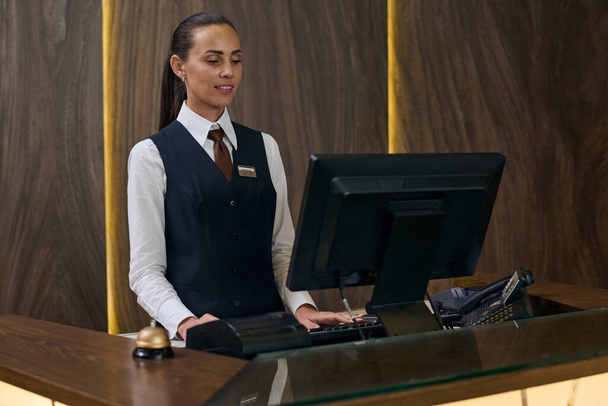Jonge receptioniste in uniform die aan de computer werkt terwijl ze achter de balie in het hotel staat - Foto, afbeelding