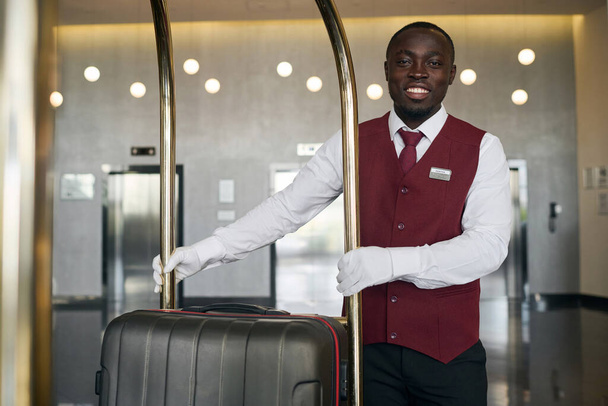 Portrait of African American doorman smiling at camera while standing with trolley for luggage - Photo, Image
