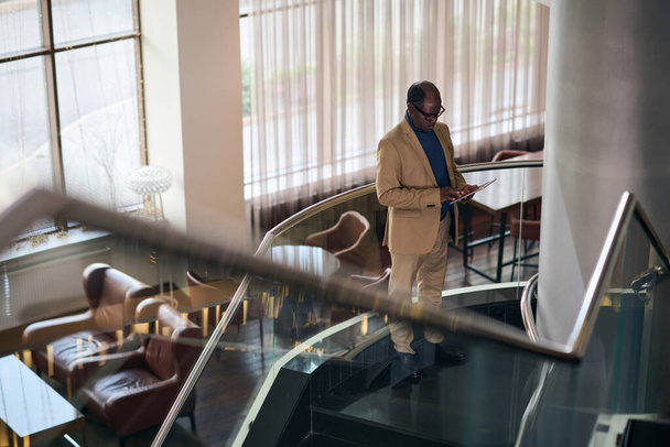 Vue grand angle de l'homme d'affaires en utilisant tablette PC tout en se tenant sur les escaliers dans le hall de l'hôtel - Photo, image
