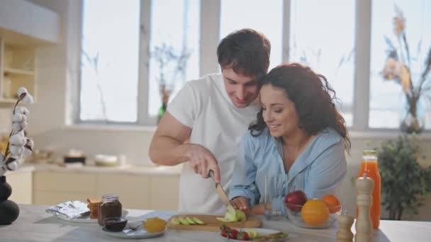 Szép házastársak vacsorát készít az egészséges reggeli a modern konyha közeli. Boldog fiatalember, aki friss almát szeletel a konyhapulton. Gyönyörű boldog nő néz főzés férj szeretettel. - Felvétel, videó