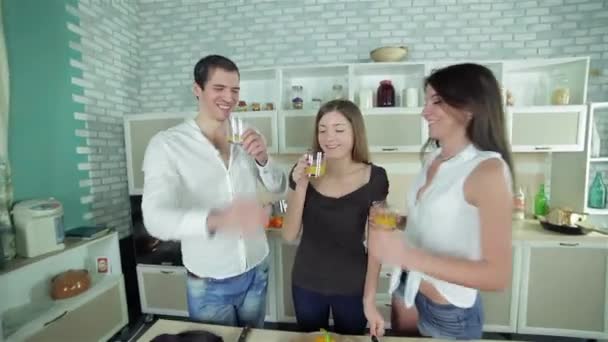 Kolme ystävää tervehtii toisiaan ja juo appelsiinimehua. Nuoret hymyilevät ystävät kokkaavat ruokaa keittiössä leikatessaan vihanneksia. Ruoanlaitto yhdessä
. - Materiaali, video