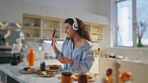Zpívající žena připravuje snídani ve sluchátkách v pohodlné domácí kuchyni zblízka. Uvolněné radostné děvče poslouchající oblíbenou píseň ve sluchátkách vaření sám. Atraktivní šťastná dáma dělá chutné přípitky. - Záběry, video