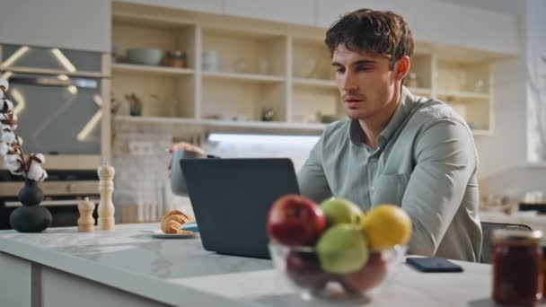Красивий фрілансер, який працює на ноутбуці, сидить на кухонному столі крупним планом. Молодий розумний чоловік дивиться на екран комп'ютера, п'є каву вдома. Серйозний працівник, який вирішує бізнес-задачі, дистанційно використовує блокнот - Кадри, відео