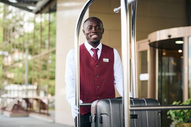 Portret afroamerykańskiego odźwiernego stojącego na zewnątrz przy wózku z bagażem i uśmiechniętym do kamery - Zdjęcie, obraz