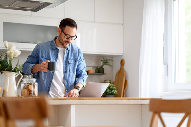 Сосредоточенный бизнесмен держит чашку кофе и читает электронную почту за ноутбуком на кухонном столе дома - Фото, изображение