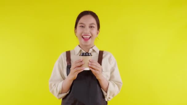 Retrato de una joven barista asiática usando delantal sosteniendo taza de café sobre fondo amarillo, camarera o emprendedora sosteniendo café con excitada, pequeña empresa o startup, camarera de cafetería. - Imágenes, Vídeo