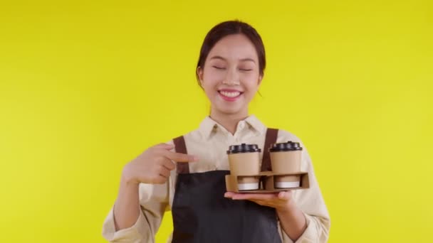 Retrato joven barista asiática mujer usando delantal sosteniendo taza de café y presentando sobre fondo amarillo, camarera o emprendedor alegre y mostrando, pequeña empresa o puesta en marcha. - Metraje, vídeo
