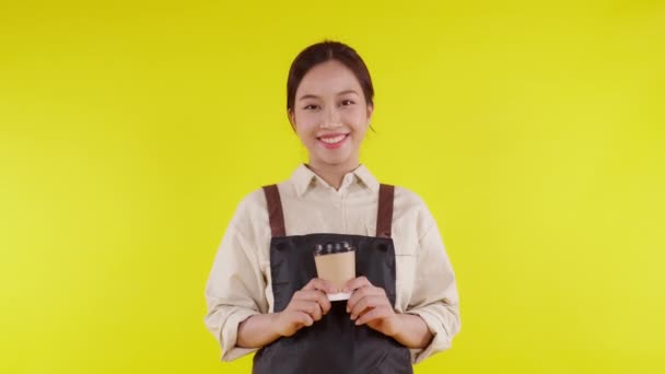 Retrato de una joven barista asiática usando delantal dando taza de café sobre fondo amarillo, camarera o emprendedora sosteniendo café con excitada, pequeña empresa o startup, camarera de cafetería. - Imágenes, Vídeo