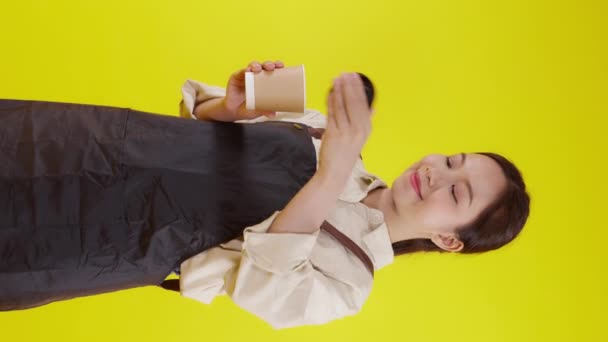 Retrato de una joven barista asiática vestida de delantal con olor a taza de café sobre fondo amarillo, camarera o emprendedora sosteniendo café con excitada, pequeña empresa o startup, camarera de cafetería. - Metraje, vídeo