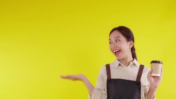 Retrato de una joven barista asiática usando delantal sosteniendo taza de café y presentando sobre fondo amarillo, camarera o emprendedora alegre y mostrando, pequeña empresa o startup, camarera de cafetería. - Imágenes, Vídeo