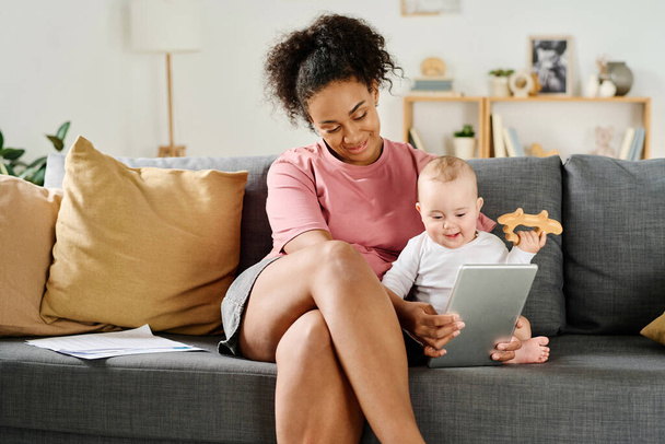 赤ちゃんと遊んでいる間ソファーに座ってタブレットのPCを使用している若い母親 - 写真・画像