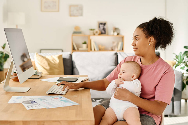 妊娠休暇中に赤ちゃんとテーブルに座ってコンピュータにタイピングする若い女性 - 写真・画像