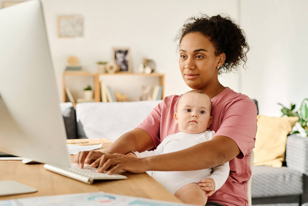 モニターの前にテーブルで赤ちゃんと座っている間,コンピュータでオンラインで働く母 - 写真・画像