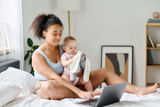 Giovane mamma seduta sul letto con il suo bambino e utilizzando il computer portatile per il suo lavoro online - Foto, immagini