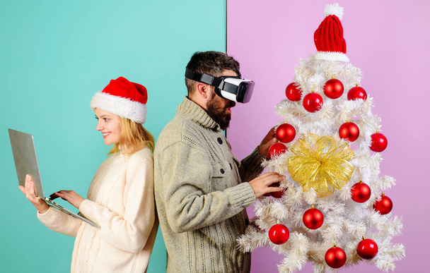 クリスマスカップル。 VRメガネの男と笑顔の女性は,自宅のクリスマスツリー近くのラップトップでサンタの帽子をかぶった. 3Dテクノロジーとエンターテイメント。 冬季休暇について。 クリスマスと新年のお祝い - 写真・画像