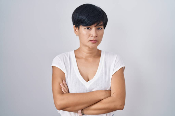 Kısa saçlı genç Asyalı kadın izole edilmiş arka planda duruyor şüpheci ve gergin, yüzünde çapraz kollu tasvip etmeyen bir ifade. negatif kişi.  - Fotoğraf, Görsel