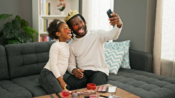 アフリカ系アメリカ人の父親と娘は王冠とバギーの目パッドを着用して自宅でスマートフォンでセルフィーを作ります - 写真・画像