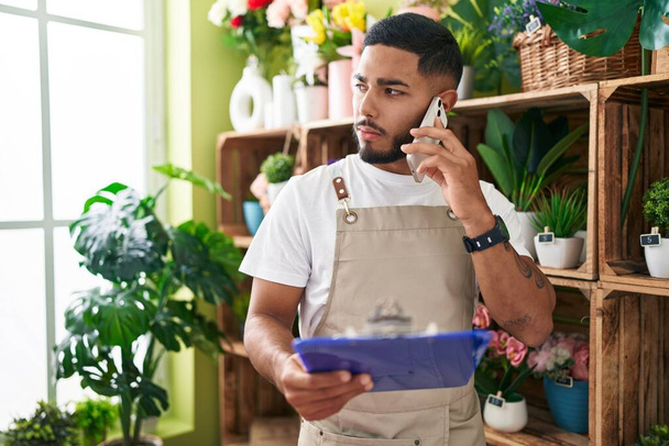 Young latin man florist talking on smartphone reading document at flower shop - Photo, Image