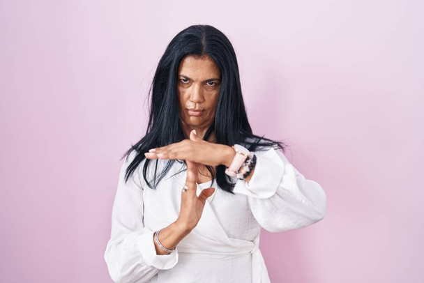 volwassen latino vrouw staande over roze achtergrond doen tijd uit gebaar met de handen, gefrustreerd en ernstig gezicht  - Foto, afbeelding