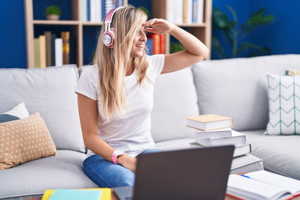 Νεαρή ξανθιά γυναίκα σπουδάζει χρησιμοποιώντας φορητό υπολογιστή στο σπίτι πολύ χαρούμενος και χαμογελαστός κοιτάζοντας μακριά με το χέρι πάνω από το κεφάλι. Αναζήτηση έννοιας.  - Φωτογραφία, εικόνα