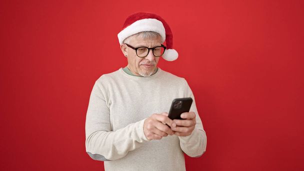 Középkorú ősz hajú férfi visel karácsonyi kalap segítségével okostelefon elszigetelt piros háttér - Fotó, kép