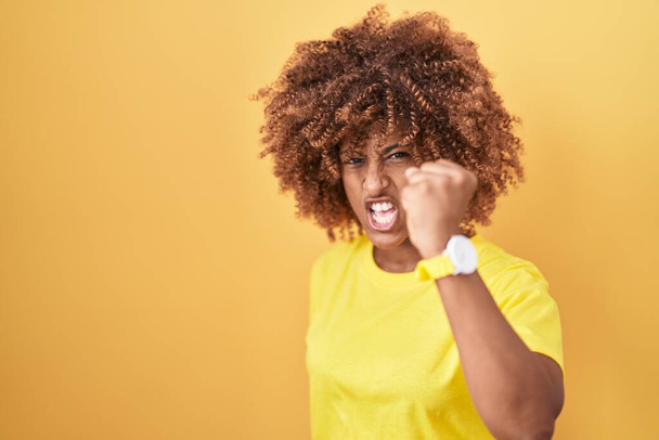 Fiatal spanyol nő göndör haj áll a sárga háttér dühös és őrült emelő ököl frusztrált és dühös, miközben kiabál a harag. düh és agresszív koncepció.  - Fotó, kép